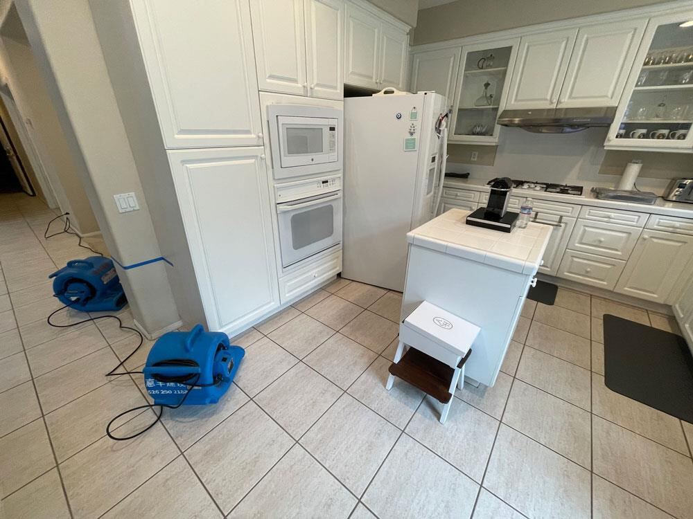 Air movers drying a kitchen floor next to white cabinets after a water leak
